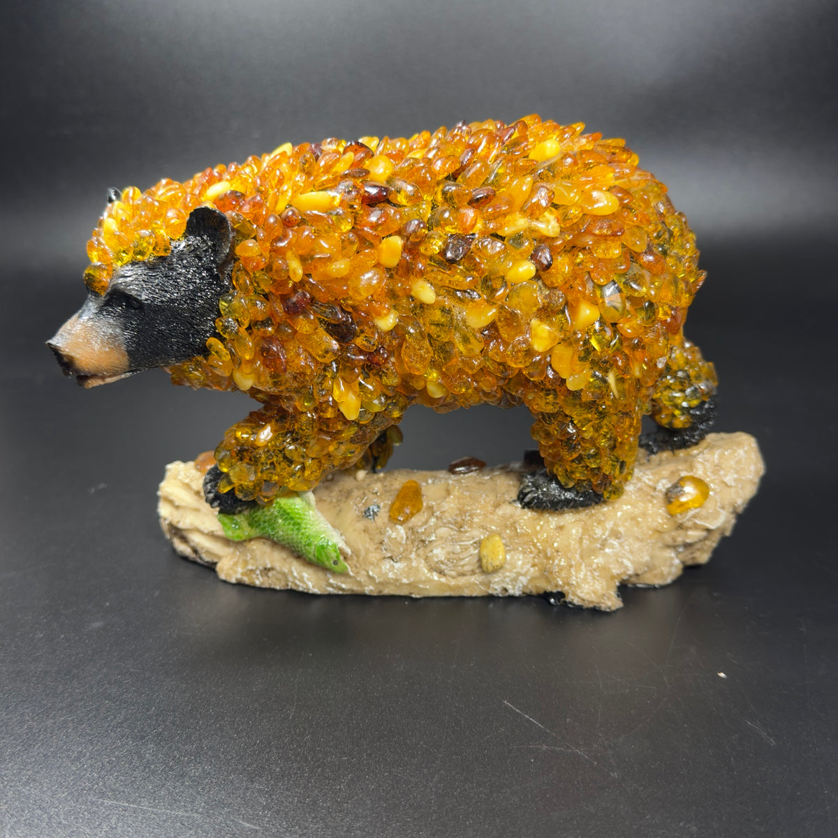 Original photograph of KIZIMA® black bear sculpture with golden Baltic amber mosaic coat, standing on a textured log beside a green fish, dark studio background