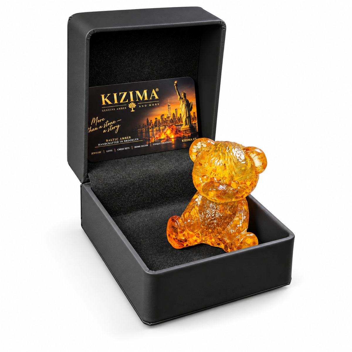 Golden amber teddy bear inside an open black KIZIMA gift box with a branded card and dark lining, photographed on white.