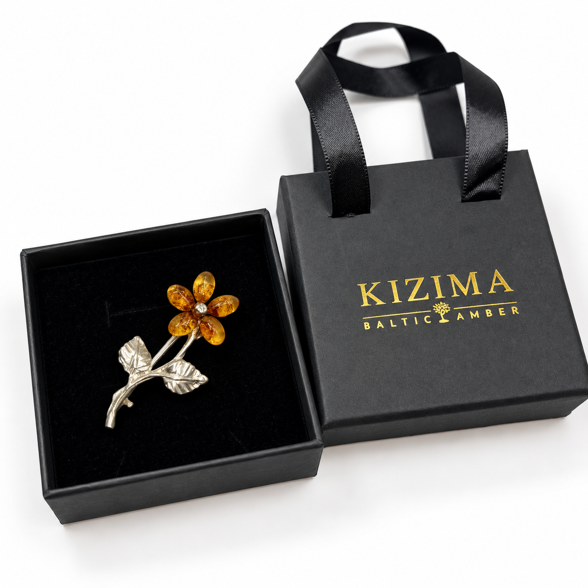 Five-petal cognac amber flower brooch in a black jewelry box beside KIZIMA Baltic Amber packaging with gold logo and satin ribbon handles.