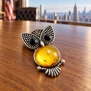 KIZIMA amber owl brooch 20N0120-42, macro office still-life resting face-up on an oak desk with a softly blurred Manhattan office background.