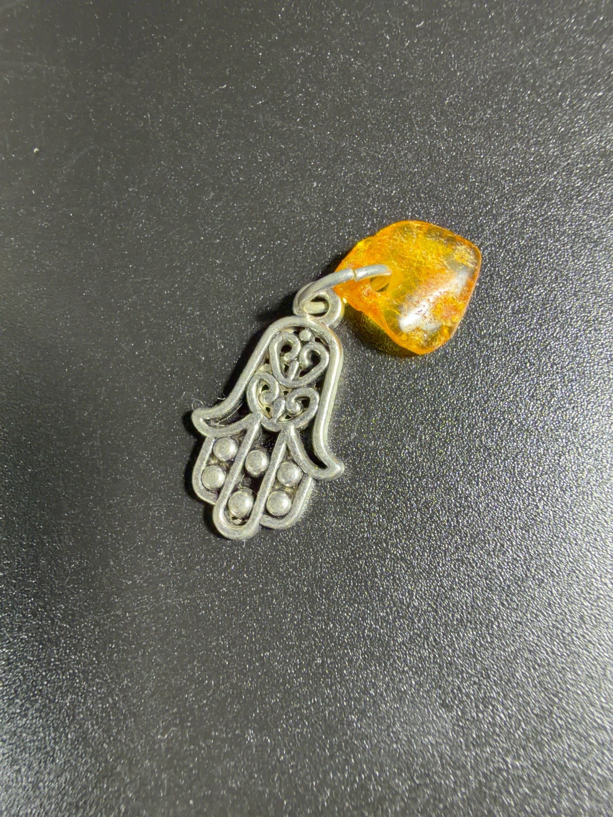Original product photograph of the KIZIMA metal Hamsa amulet with natural Baltic amber on its real tabletop background.
