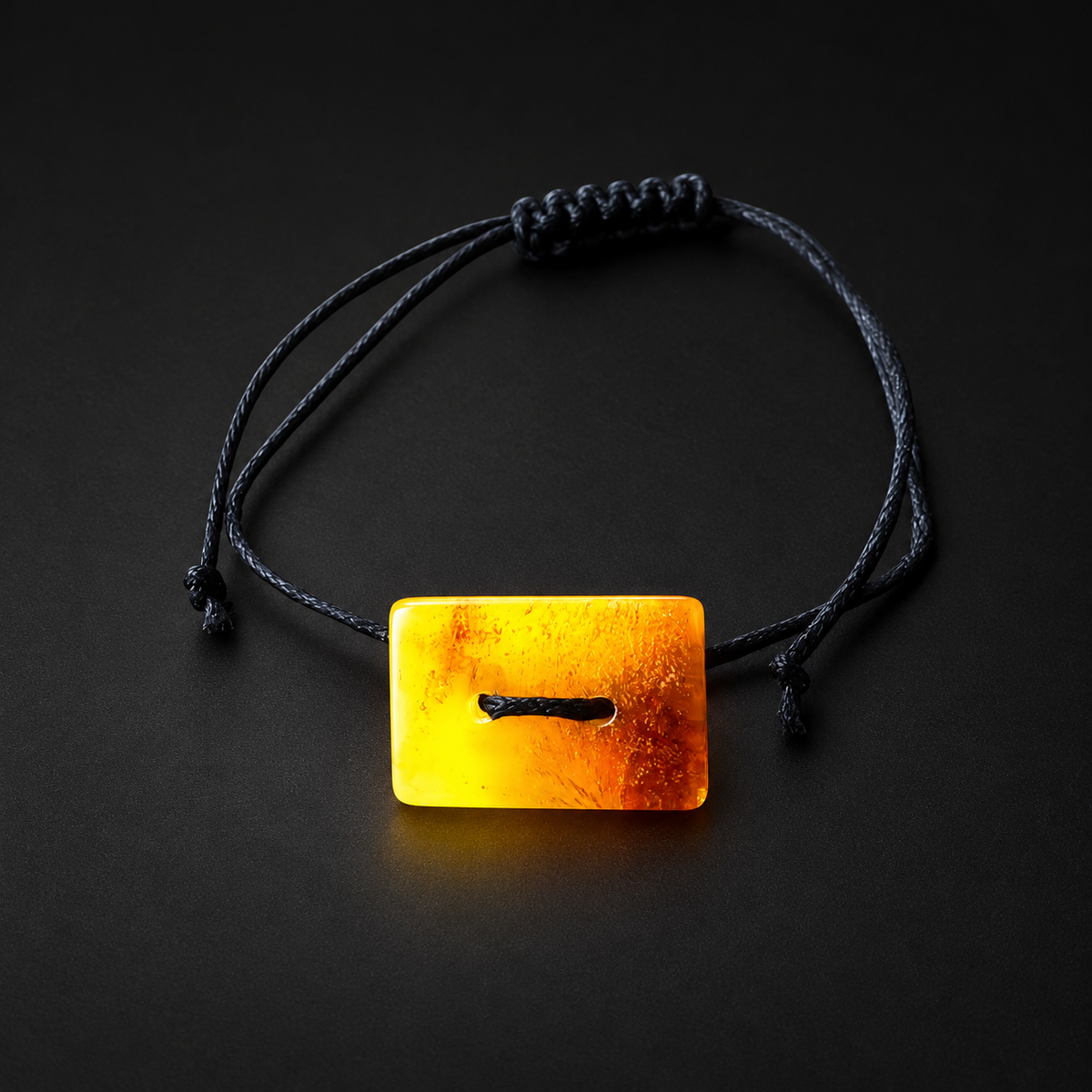 Rectangular honey amber button and black cord of KIZIMA 3G0125q1.81 bracelet against a black studio background
