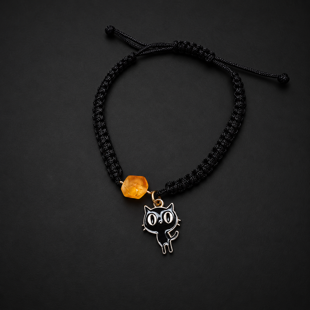 KIZIMA honey amber bracelet with a black enamel cat charm and black macrame band on a dark studio surface