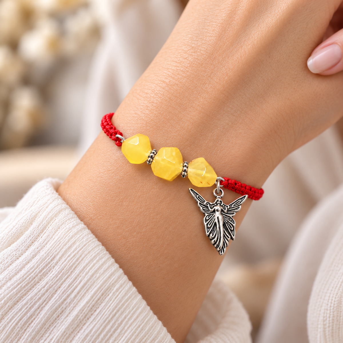 Red cord bracelet with yellow amber stones and an angel charm worn on a woman’s wrist beside an ivory sleeve.