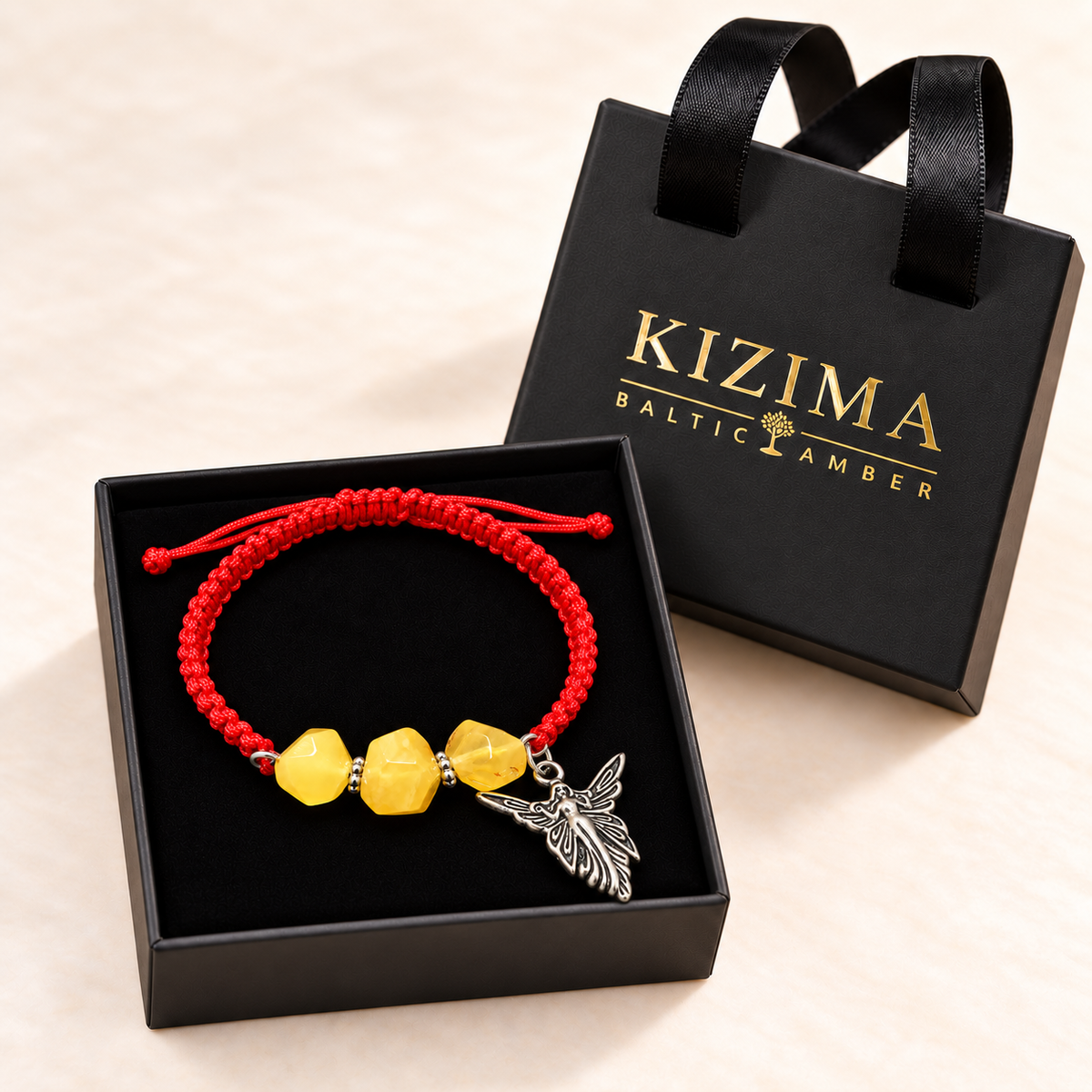 Red macrame yellow amber angel bracelet in a clean black velvet-lined KIZIMA gift box beside its handled gold-logo lid.