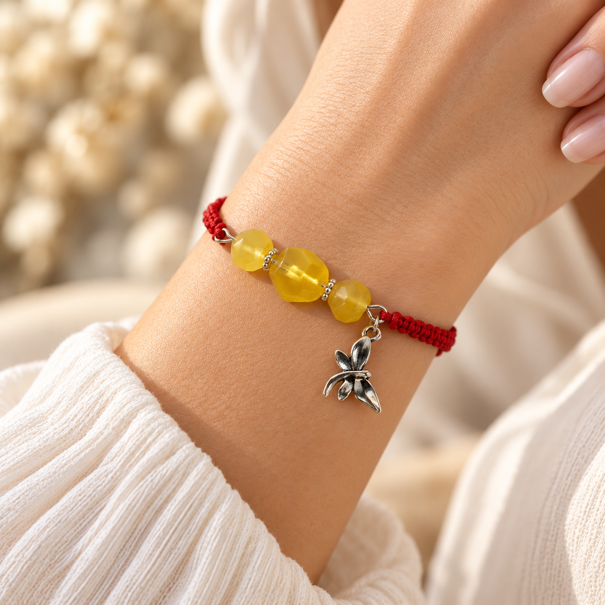 Red cord and yellow amber bracelet shown on a woman's wrist, with its silver dragonfly charm beside the stones.