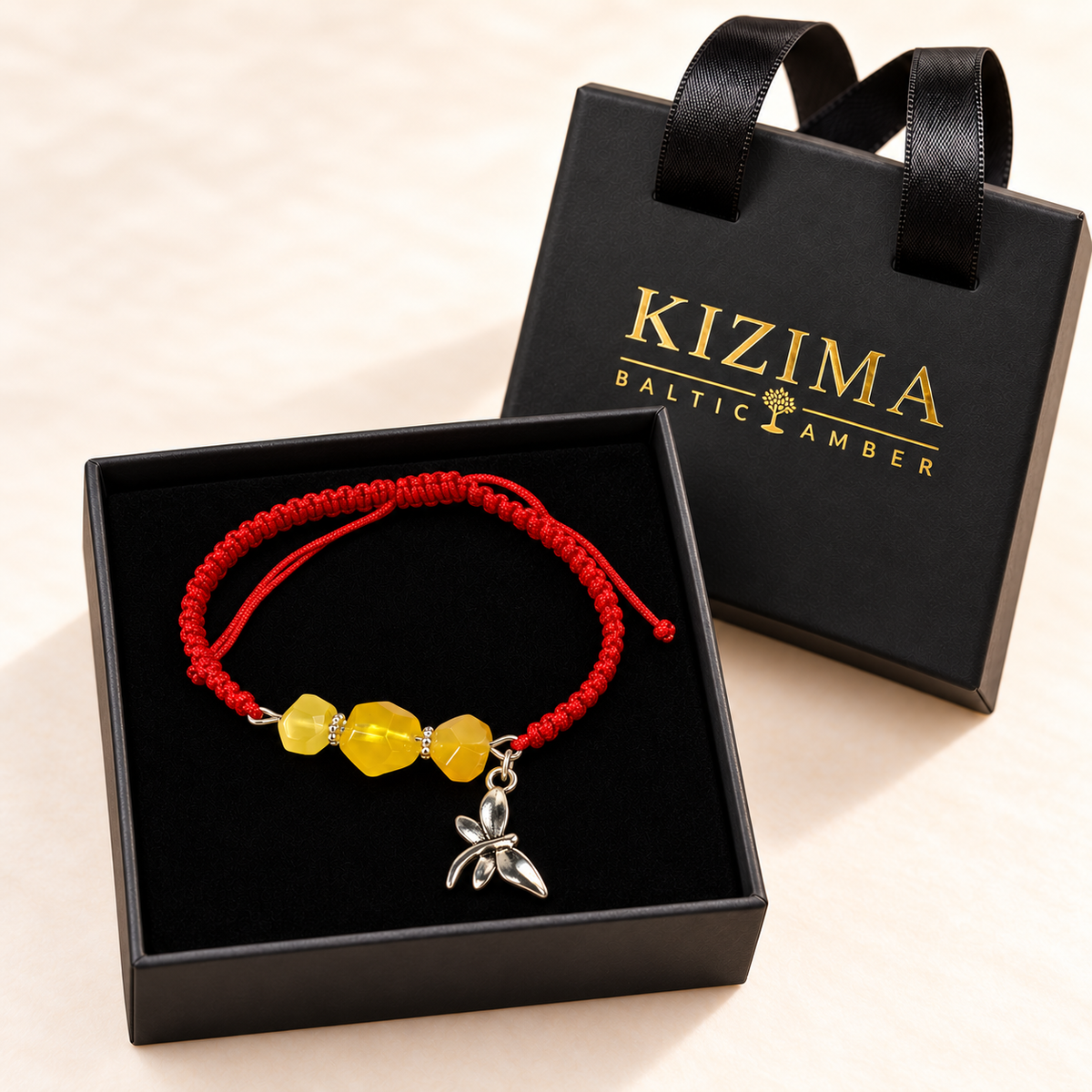 KIZIMA red cord amber bracelet with dragonfly charm arranged inside the black branded gift box.