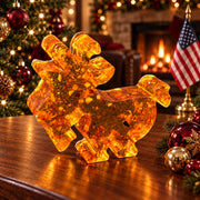 Small flat amber goat with curved horns and a beard on a walnut tabletop beside red and gold ornaments, with a Christmas tree, fireplace and American flag in the background.