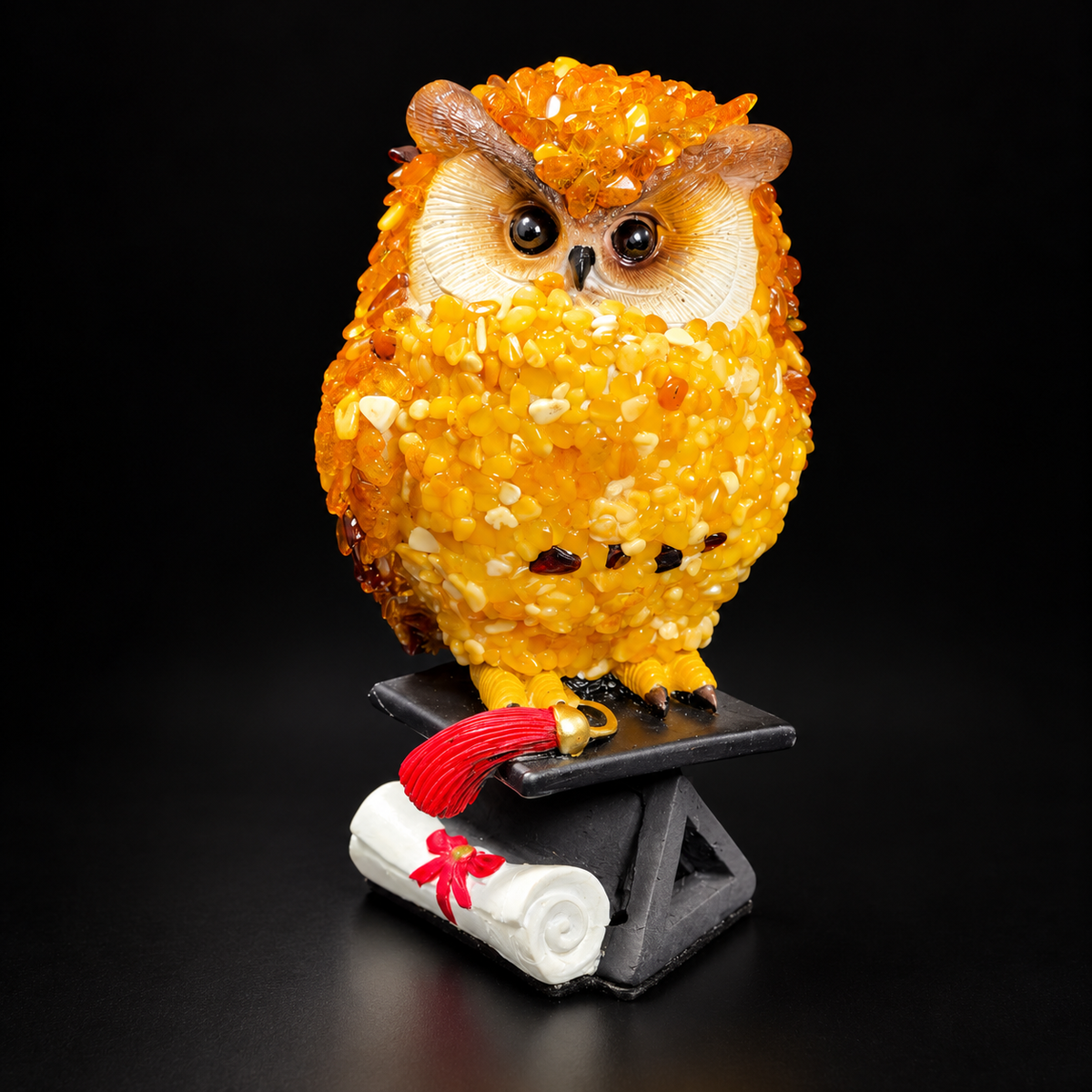 Front view of the golden Baltic amber mosaic owl with red tassel and rolled diploma on a black studio background.