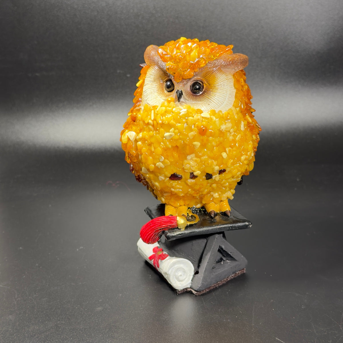 Yellow Baltic amber owl standing on a black graduation cap with a red tassel and a white diploma, front three-quarter view.