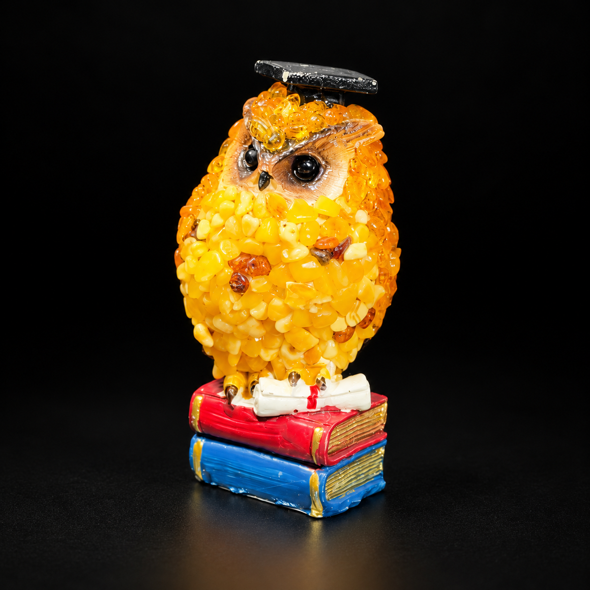 Golden amber owl in a black graduation cap with a rolled diploma and red and blue books on a black studio background.