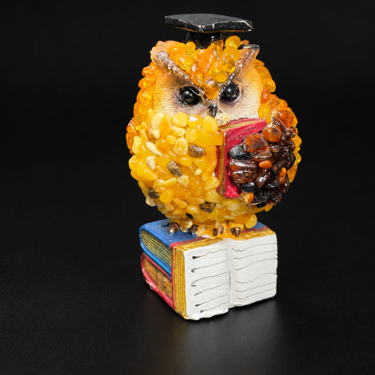 Amber owl in a graduation cap holding an open red book, golden and brown amber, on a black studio background.