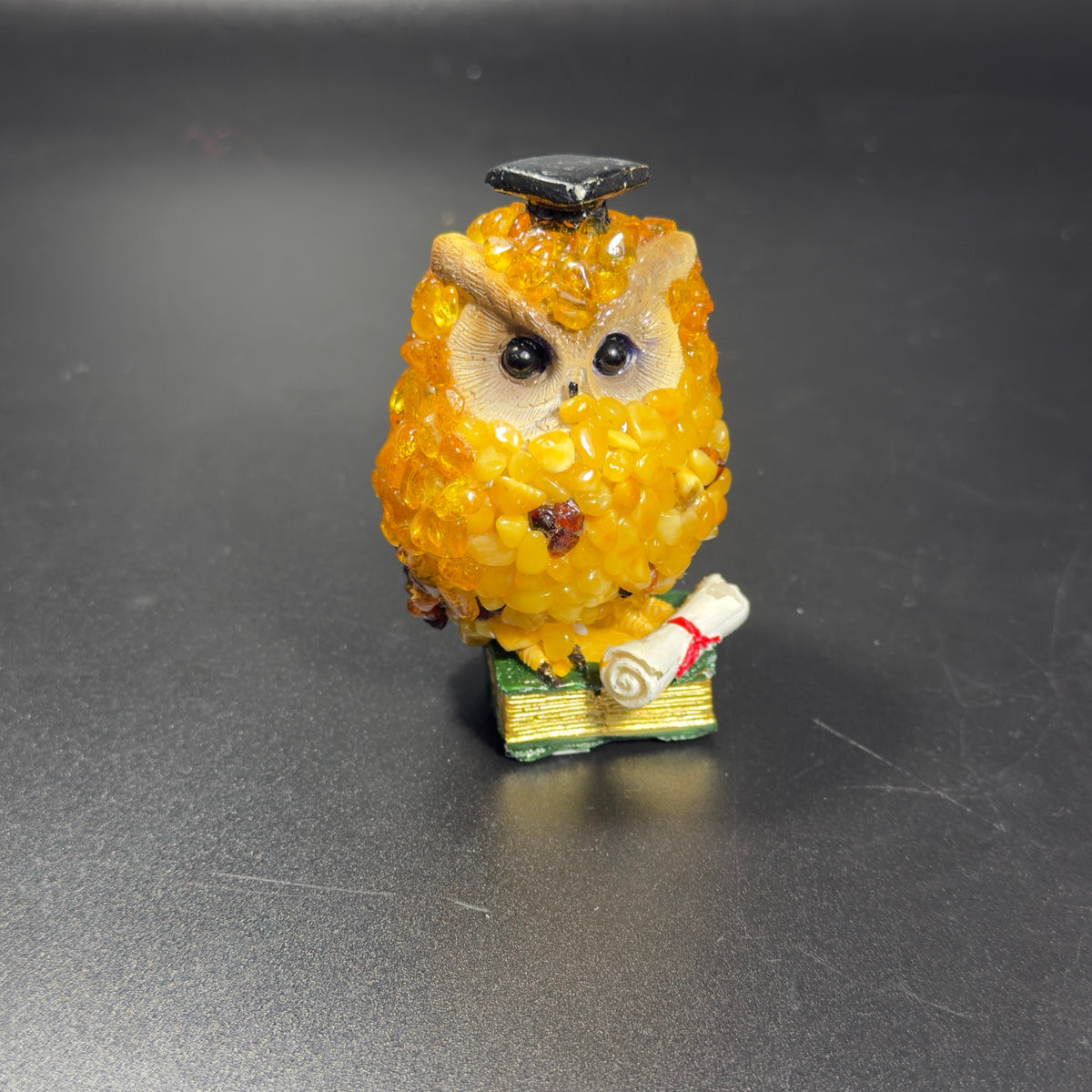 Original photo of golden-yellow graduate owl with black mortarboard, green book and white diploma tied with red ribbon