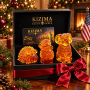 Three distinct amber sheep and ram figurines arranged from shortest to tallest inside a black KIZIMA box with red satin ribbon, against a Christmas tree, fireplace and American flag.