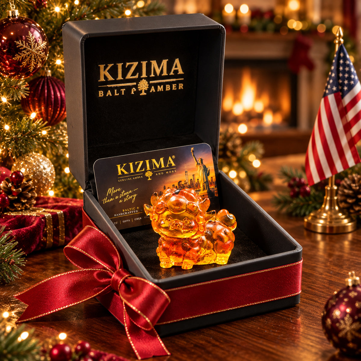 Tiny standing amber lamb with rounded fleece and an upturned tail in an open black KIZIMA gift box with a red bow, decorated tree lights and an American flag behind.