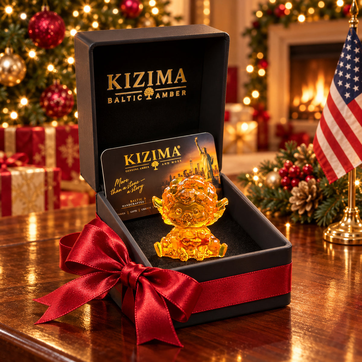 Mini seated amber sheep with short horns and outstretched hind legs in an open black KIZIMA gift box with a red bow, decorated tree lights and an American flag behind.