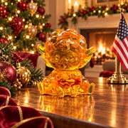 Mini seated amber sheep with short horns and outstretched hind legs displayed on walnut beside Christmas ornaments, with a glowing fireplace, decorated tree and American flag.