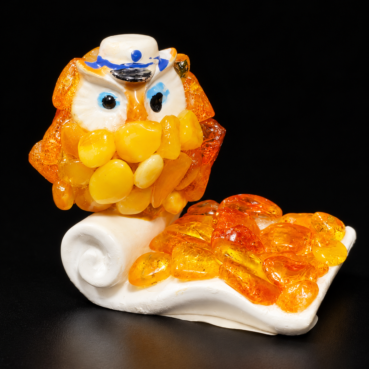 Golden-yellow owl figurine with blue eyes, white hat and curled scroll base against a black studio background