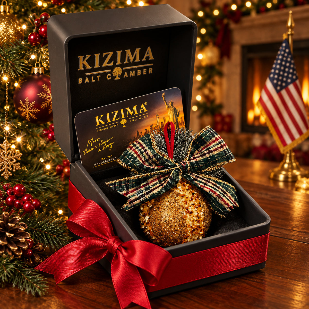 Golden textured amber ball with a green tartan bow in a black KIZIMA gift box with red satin ribbon, a Christmas tree and American flag.
