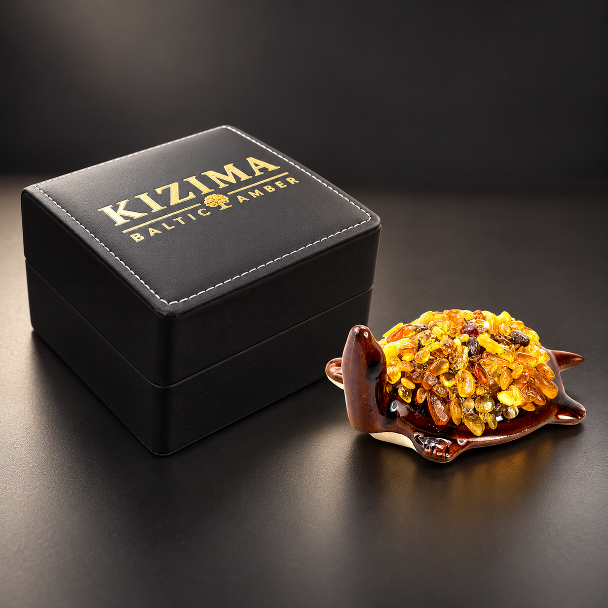 KIZIMA 10N0050q21 Baltic amber turtle figurine beside the closed black KIZIMA Baltic Amber gift box with gold branding and white stitching.