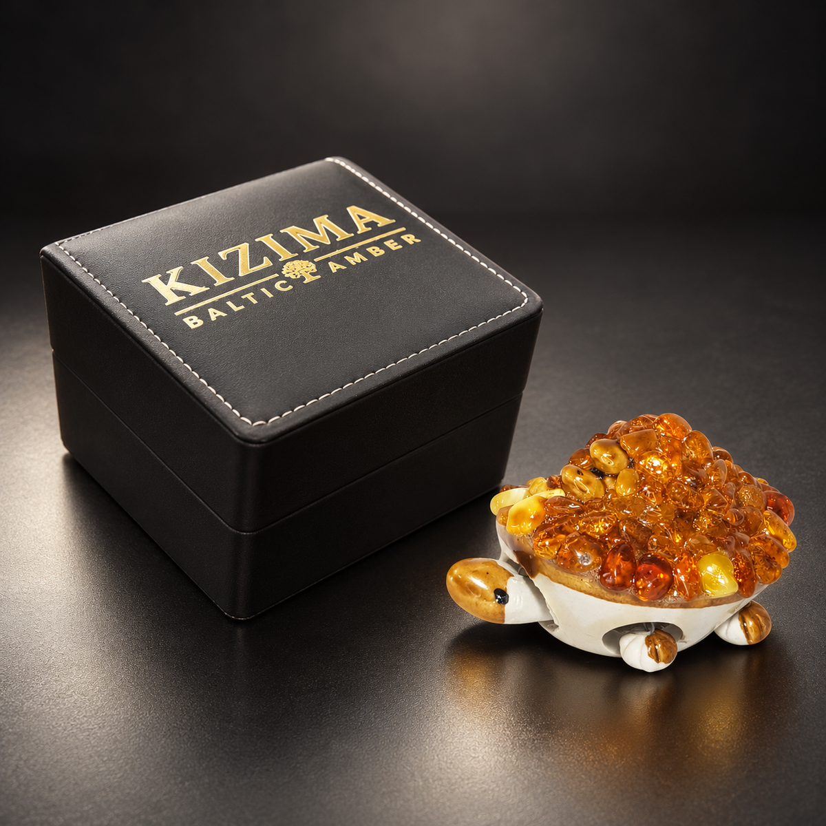 KIZIMA 10Z0100q53 Baltic amber turtle figurine beside the closed black KIZIMA Baltic Amber gift box with gold branding and white stitching.