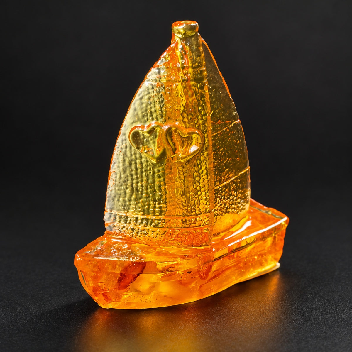 Amber sailboat with two hearts on a textured golden sail, photographed against a charcoal studio background.