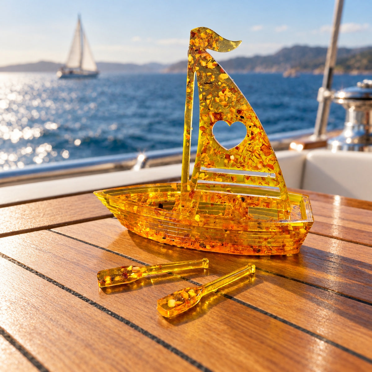 KIZIMA heart sailboat miniature and two amber oars on a teak yacht table with the blue sea behind.