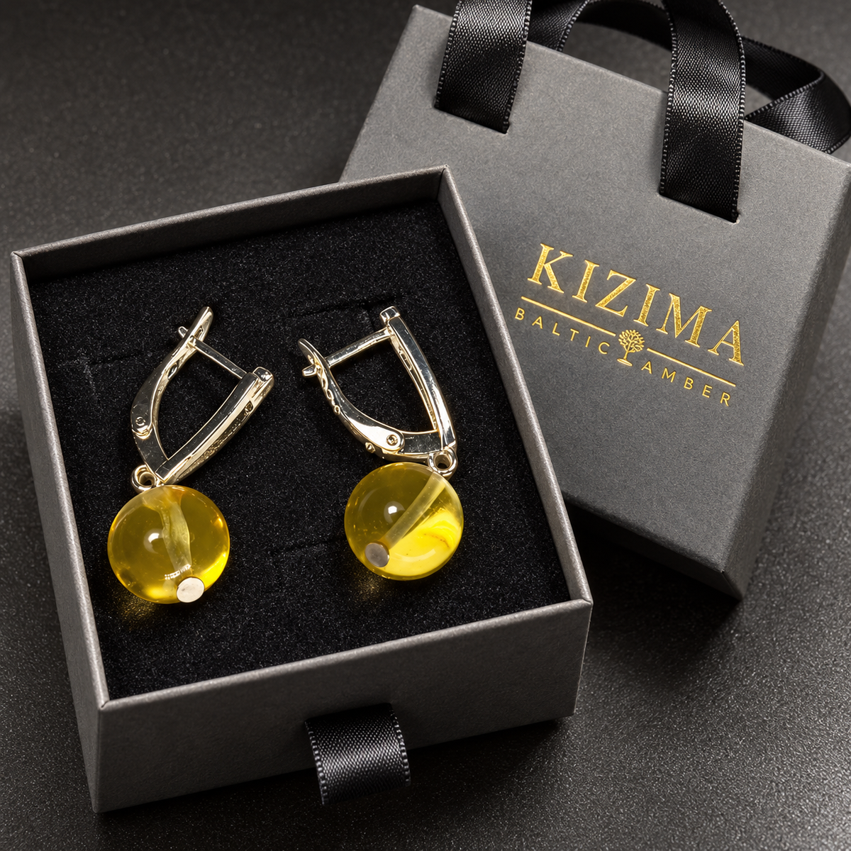 Transparent honey amber sun ball earrings in a charcoal gray KIZIMA ribbon-handle gift box with black insert.