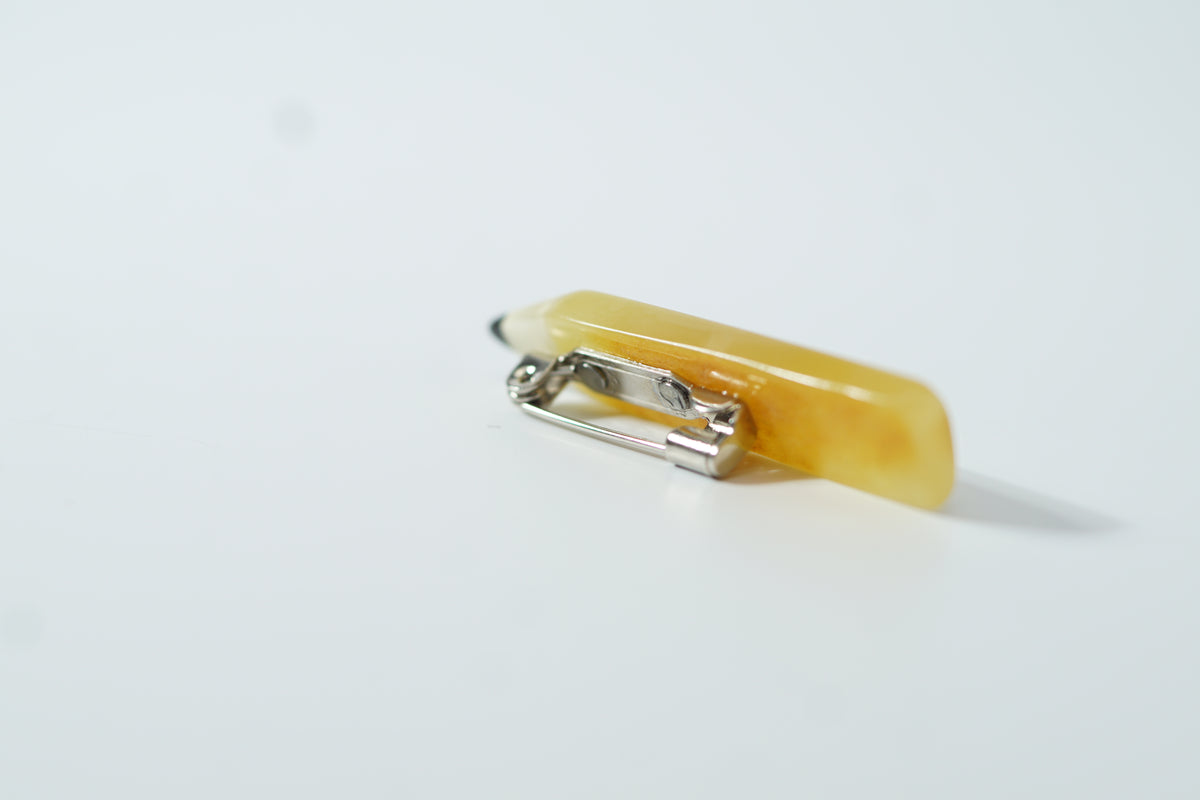 Back of a yellow amber pencil brooch with the metal clasp visible on a pale surface; a retirement gift for an educator.