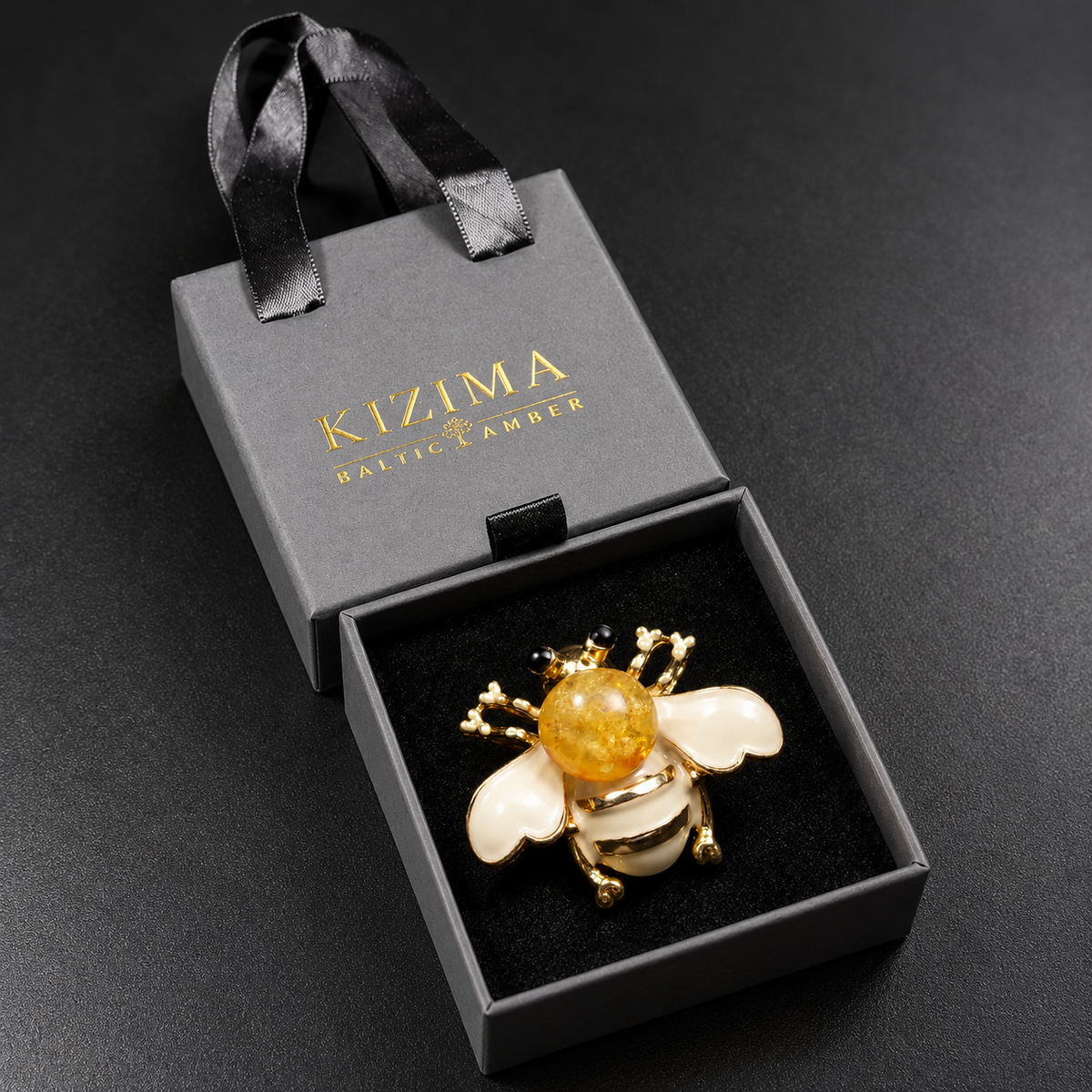 KIZIMA cream-white enamel and honey Baltic amber bee brooch 20-307Q5-3-3-2 in its charcoal-gray KIZIMA velvet-lined drawer gift box.