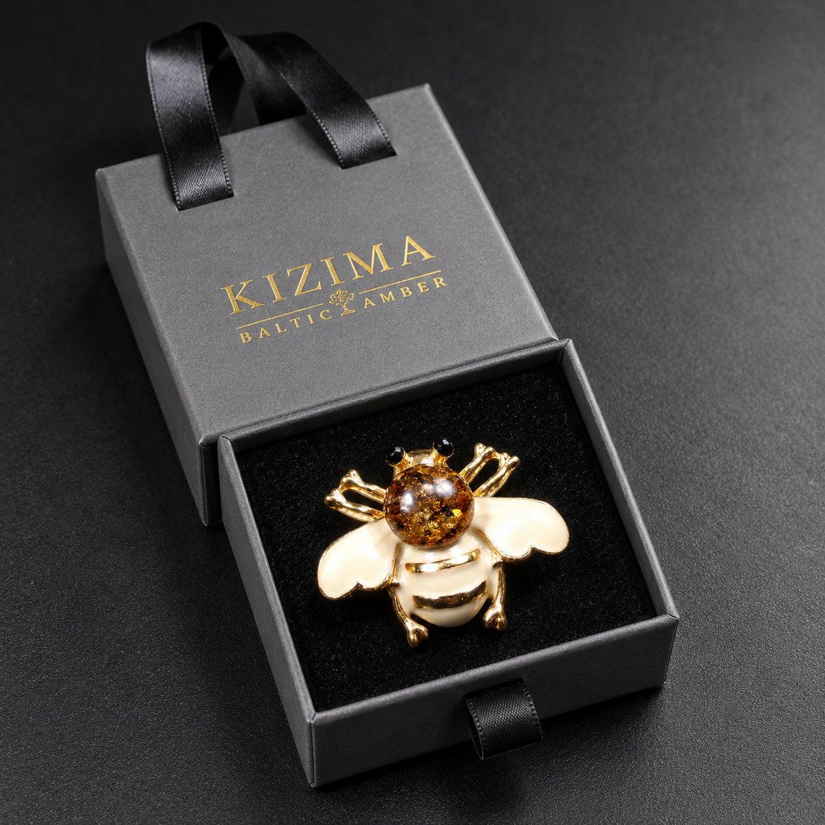 KIZIMA cream-white enamel bee brooch 20N0120-39 with round Baltic amber in its charcoal-gray KIZIMA velvet-lined drawer gift box.
