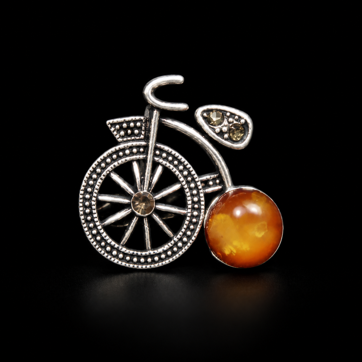 KIZIMA silver-tone bicycle brooch with cloudy honey-orange Baltic amber wheel on black studio background.