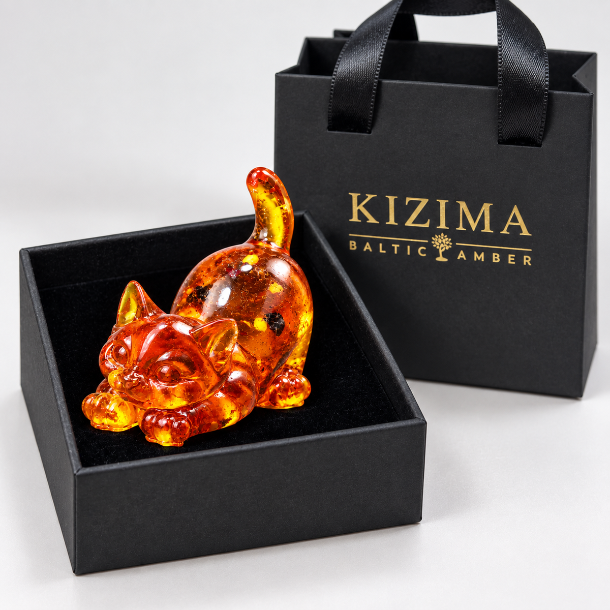 Stretching amber cat with extended paws and an upright tail with a black velvet-lined box and branded KIZIMA gift bag