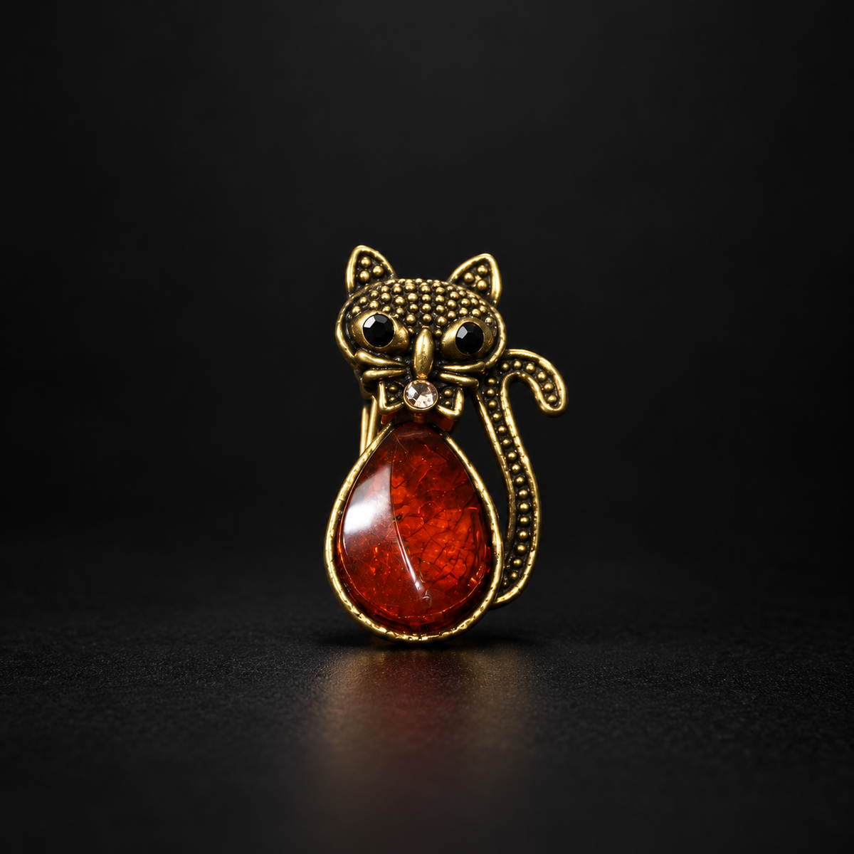 KIZIMA gold-tone cat brooch 20-206Q4-3-1-1 with reddish cognac Baltic amber belly, black eyes and a crystal bow on a black studio background.