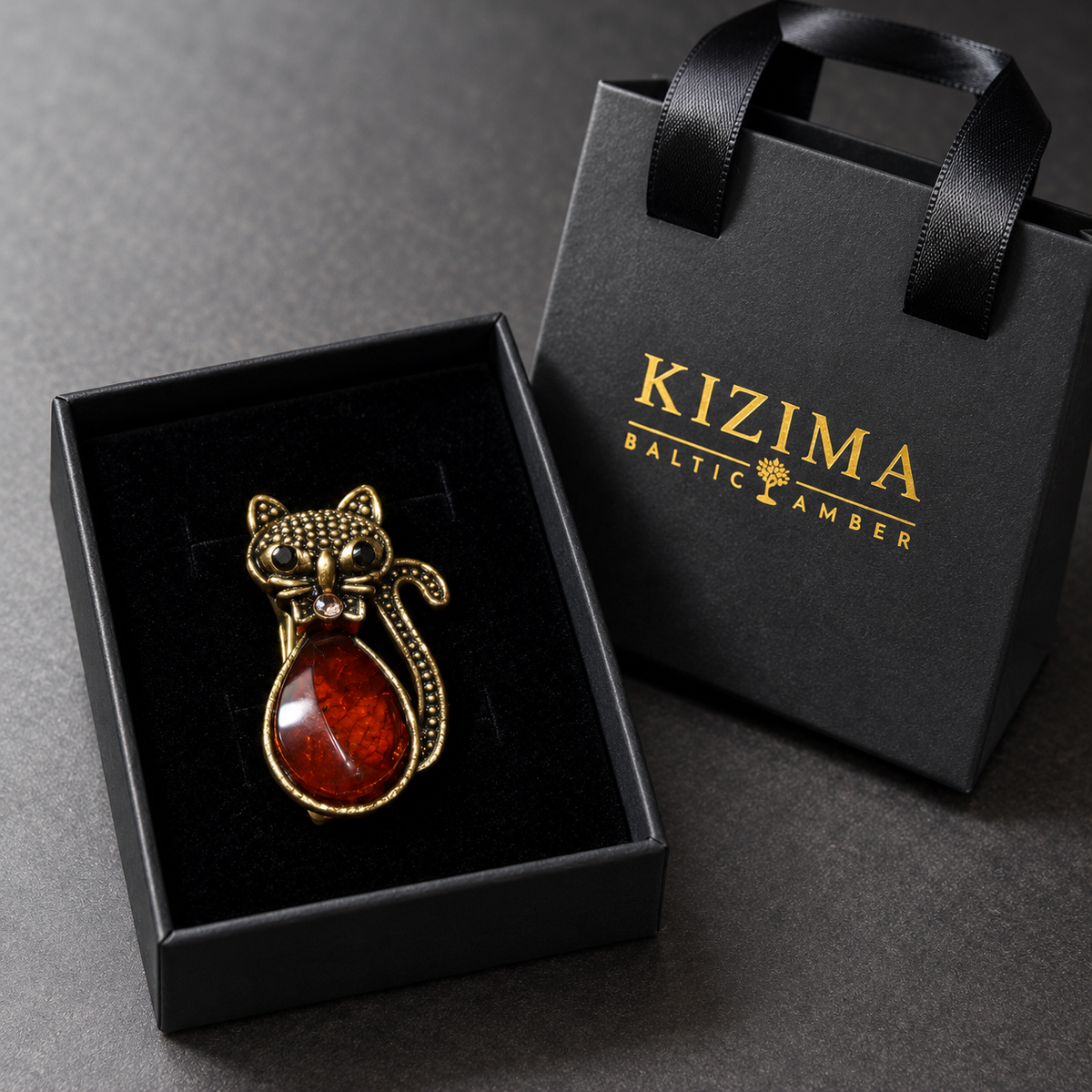 KIZIMA gold-tone cat brooch 20-206Q4-3-1-1 with reddish cognac Baltic amber belly, black eyes and a crystal bow in the real black velvet KIZIMA gift box with matching bag.