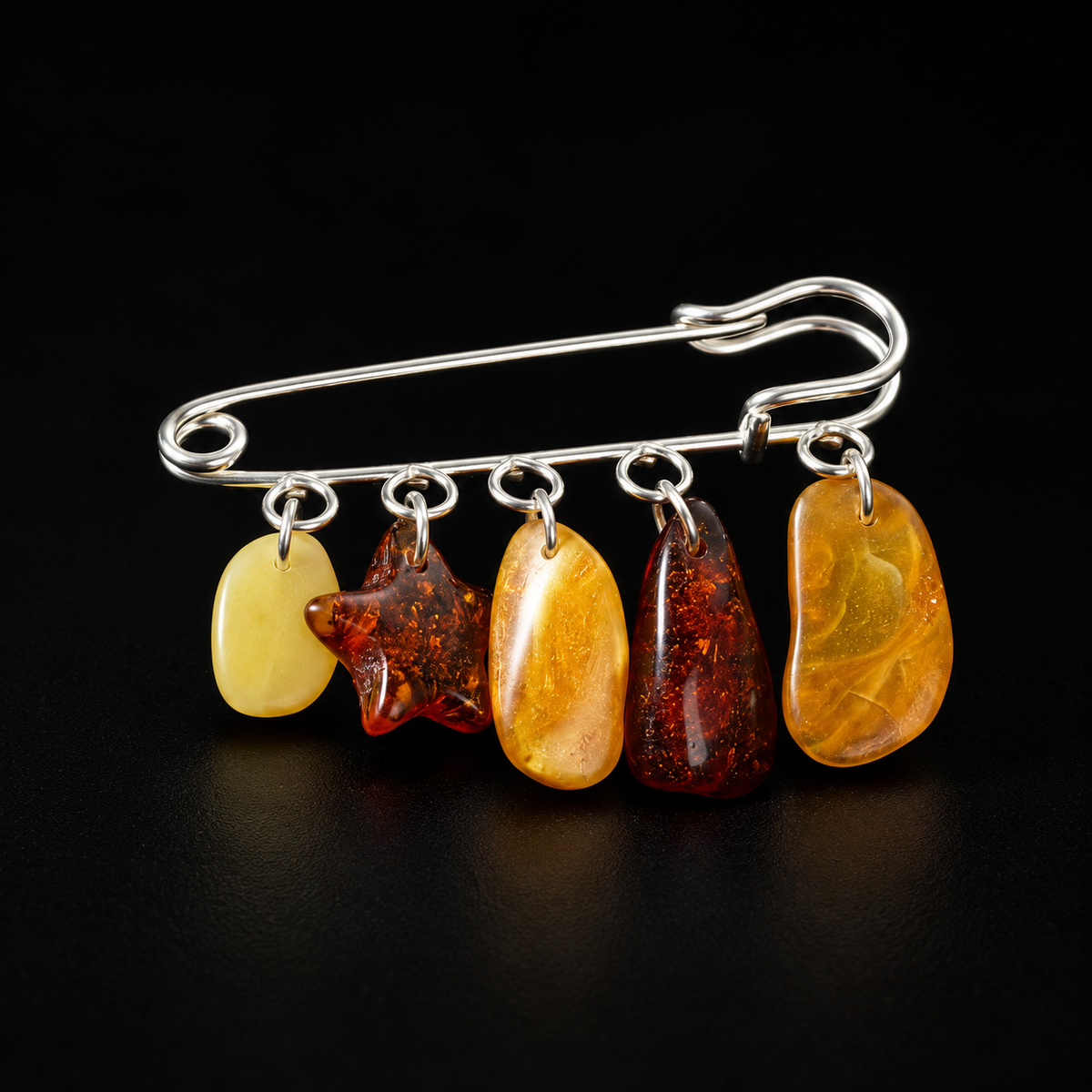 KIZIMA silver-tone safety pin brooch with five distinct yellow, golden and cognac Baltic amber charms on black studio background.