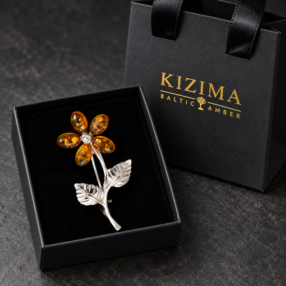 KIZIMA five-petal honey amber flower brooch with a silver-tone stem and two leaves in the real black velvet KIZIMA box beside the gold-logo gift bag.