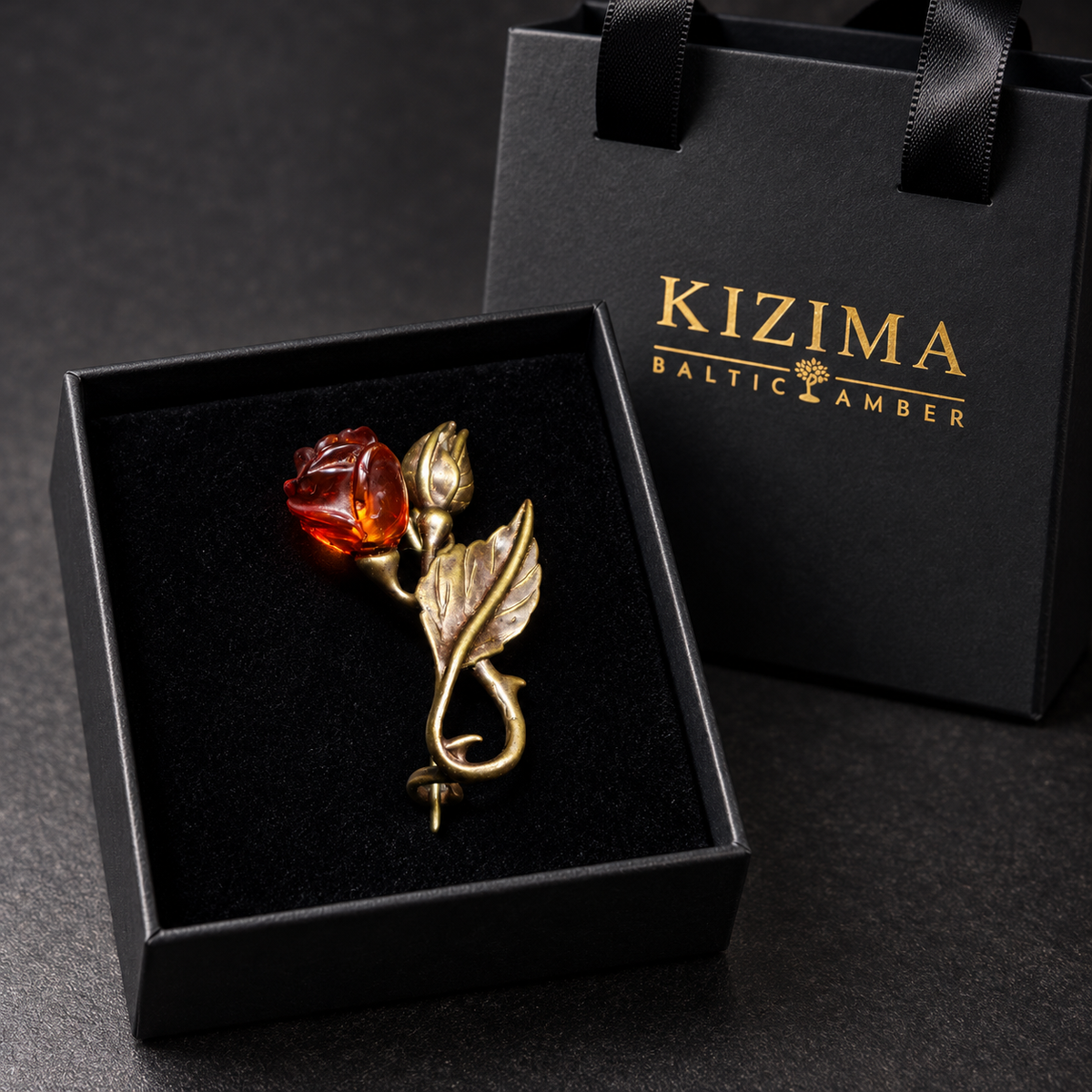 KIZIMA antique bronze-tone flower-and-bud brooch with a carved reddish cognac amber rose, metal bud and curling leaf stem in the real black velvet KIZIMA box beside the gold-logo gift bag.
