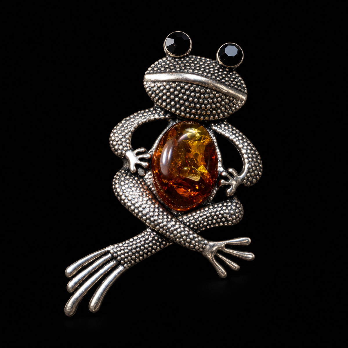 KIZIMA silver-tone seated frog brooch with black eyes and cognac Baltic amber belly on black studio background.