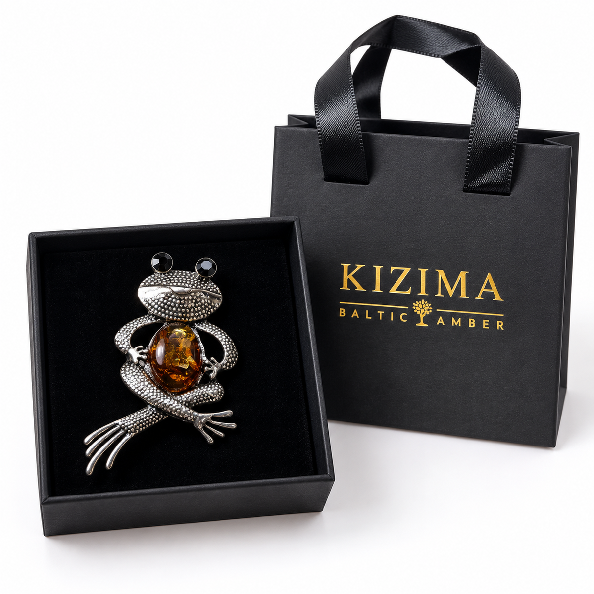 KIZIMA silver-tone seated frog brooch with black eyes and cognac Baltic amber belly in actual black KIZIMA gift packaging.