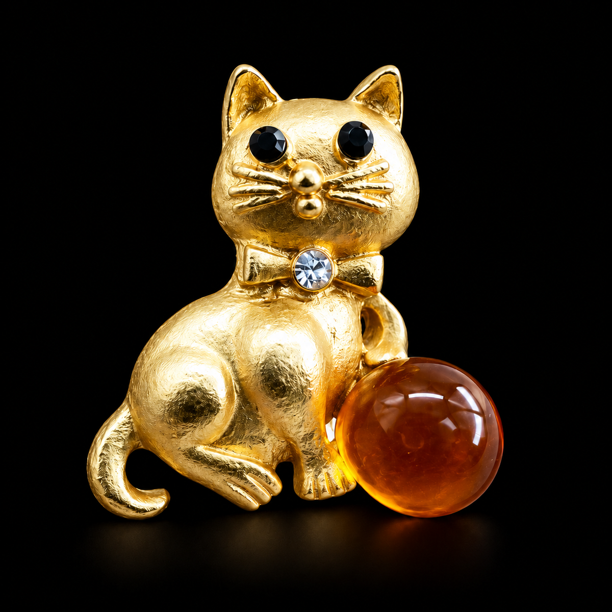 KIZIMA gold-tone seated cat brooch with black eyes, clear bowtie crystal and cognac Baltic amber sphere on black studio background.