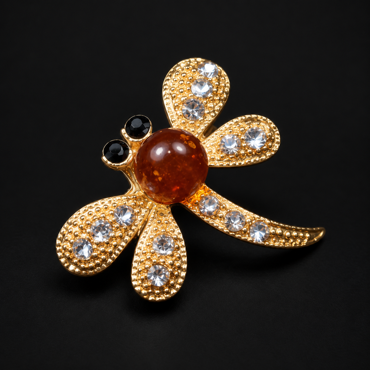 KIZIMA gold-tone dragonfly brooch with cognac amber center and clear crystals on a black background.