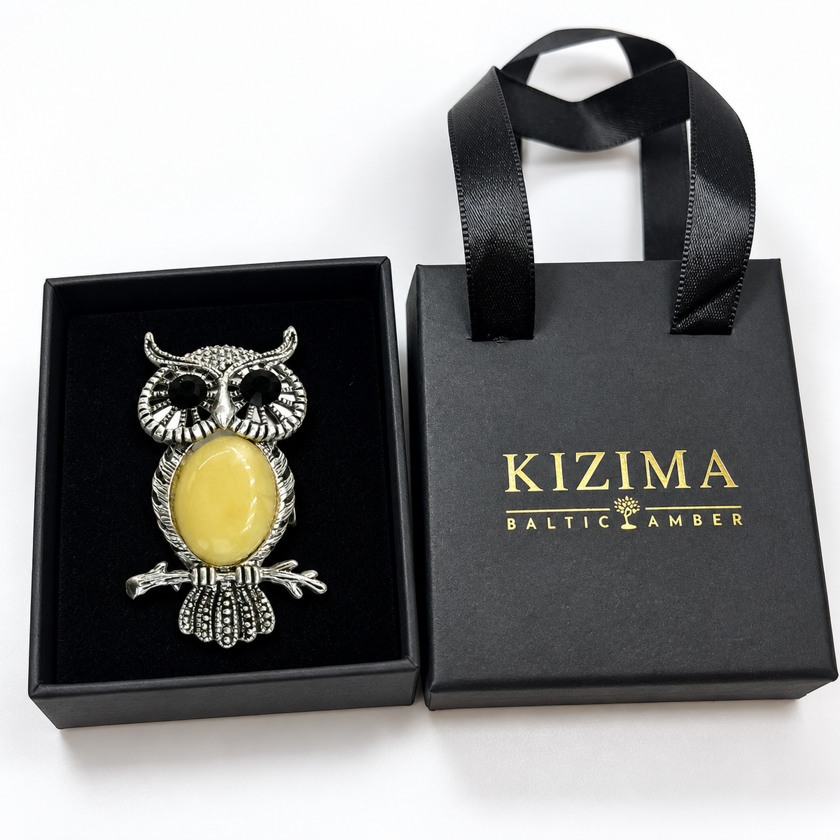 KIZIMA silver-tone owl brooch with black eyes and opaque pale-yellow Baltic amber belly in actual black KIZIMA gift packaging.