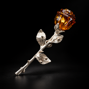KIZIMA silver-tone rose brooch with a carved cognac Baltic amber bud and three veined leaves on a black studio background.