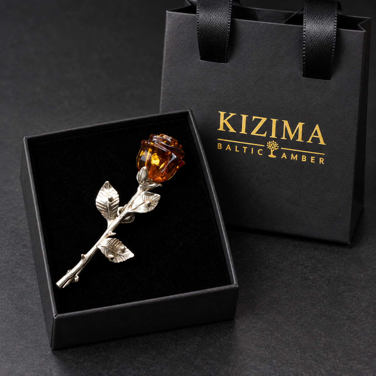 KIZIMA silver-tone rose brooch with a carved cognac Baltic amber bud and three veined leaves in the real black velvet KIZIMA box beside the gold-logo gift bag.
