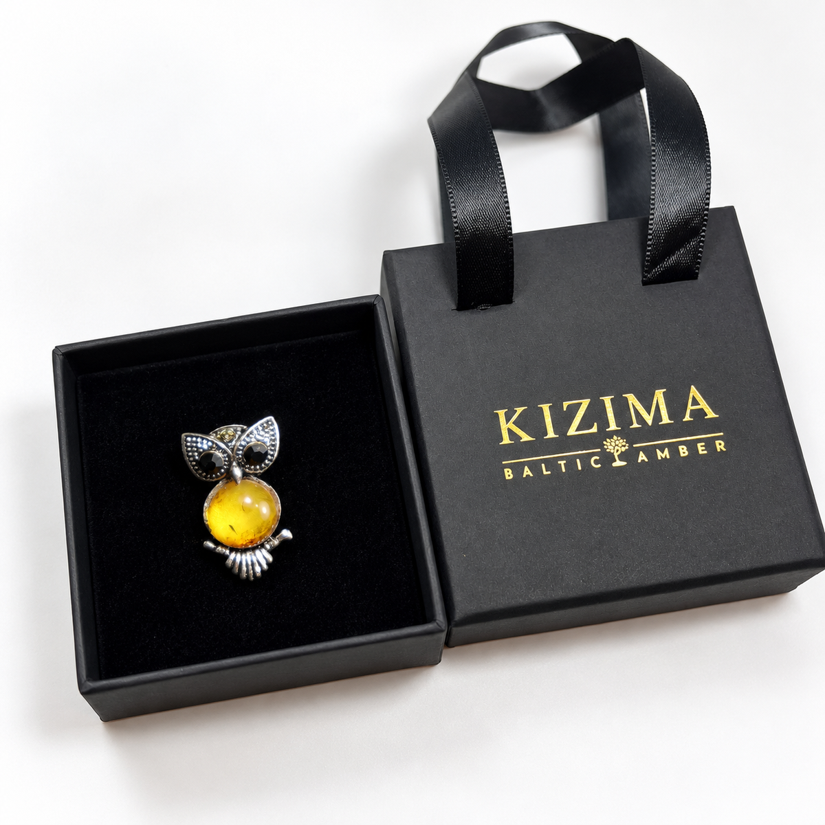 KIZIMA small silver-tone owl brooch with black eyes and honey-yellow Baltic amber belly in actual black KIZIMA gift packaging.