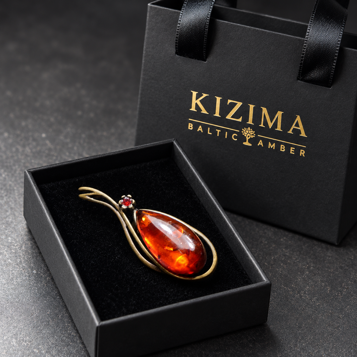 KIZIMA antique bronze-tone teardrop brooch with reddish cognac Baltic amber and a small red accent stone in the real black velvet KIZIMA box beside the gold-logo gift bag.
