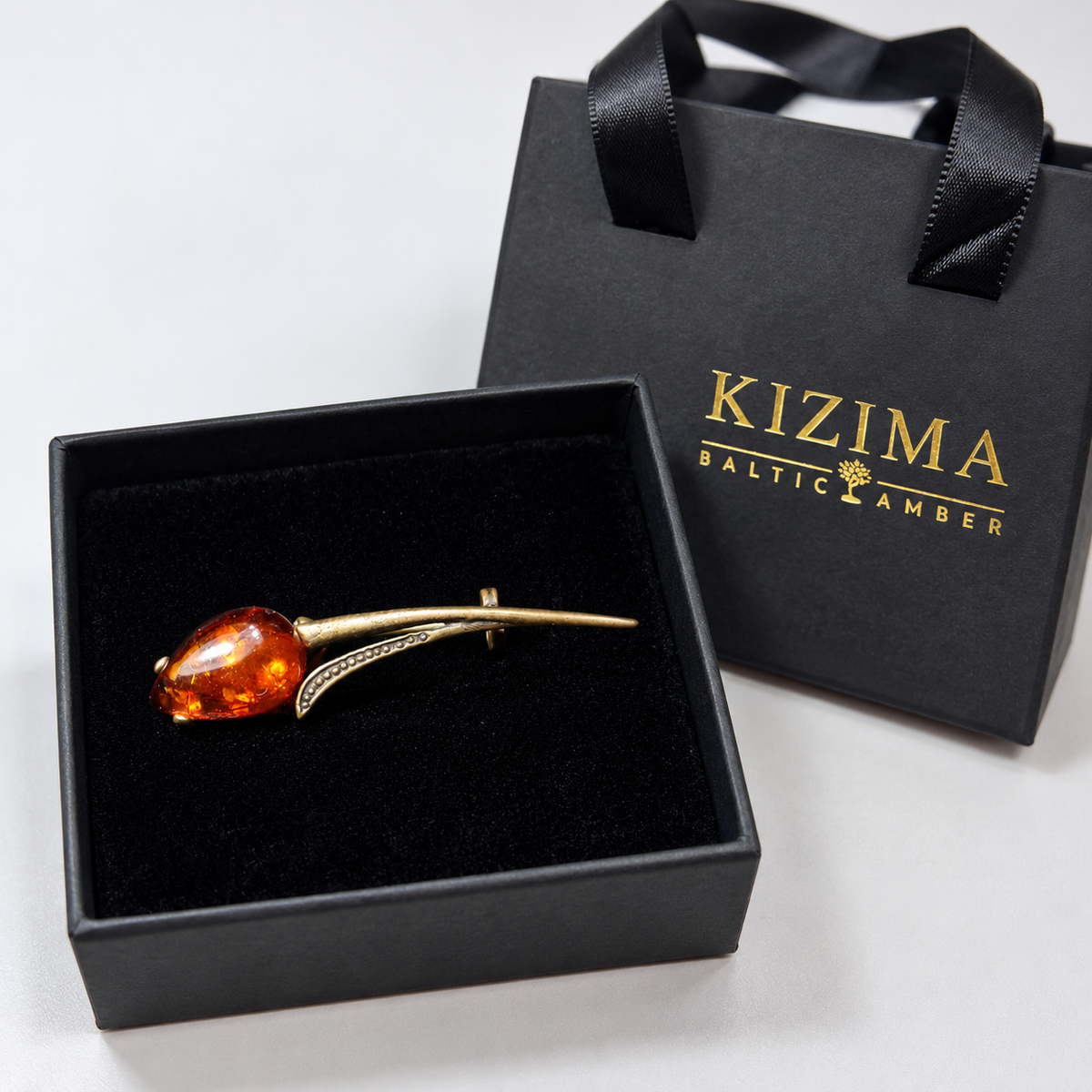 KIZIMA antique bronze-tone tulip brooch with a cognac amber teardrop bloom and long curved stem in the real black velvet KIZIMA box beside the gold-logo gift bag.