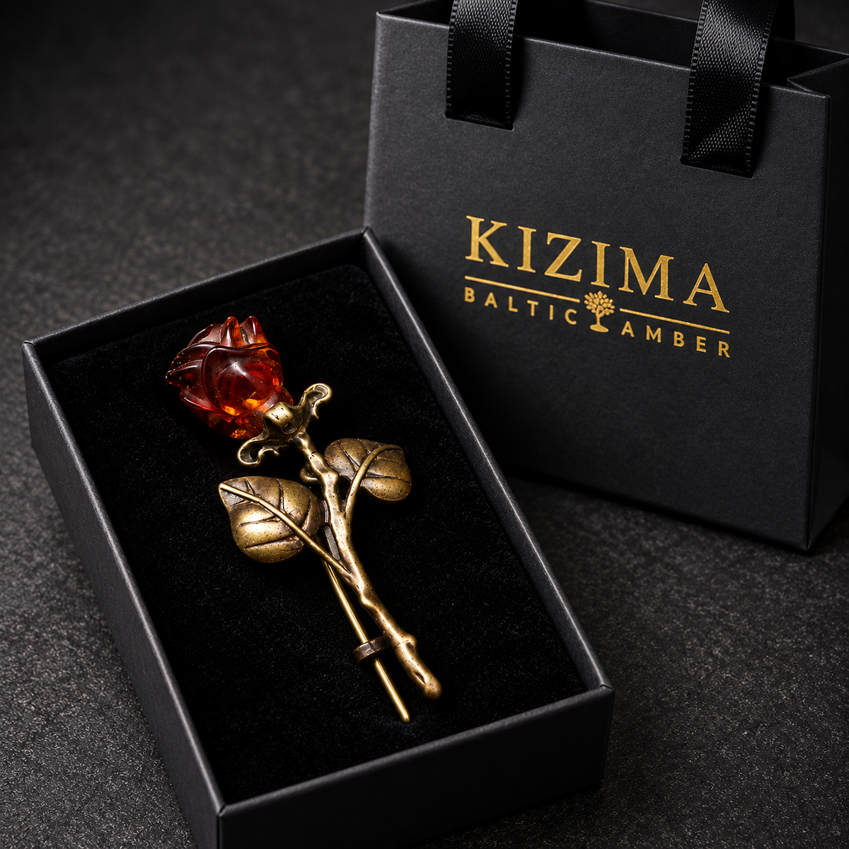 KIZIMA antique bronze-tone rose brooch with a deep cognac amber bud and two rounded leaves in the real black velvet KIZIMA box beside the gold-logo gift bag.